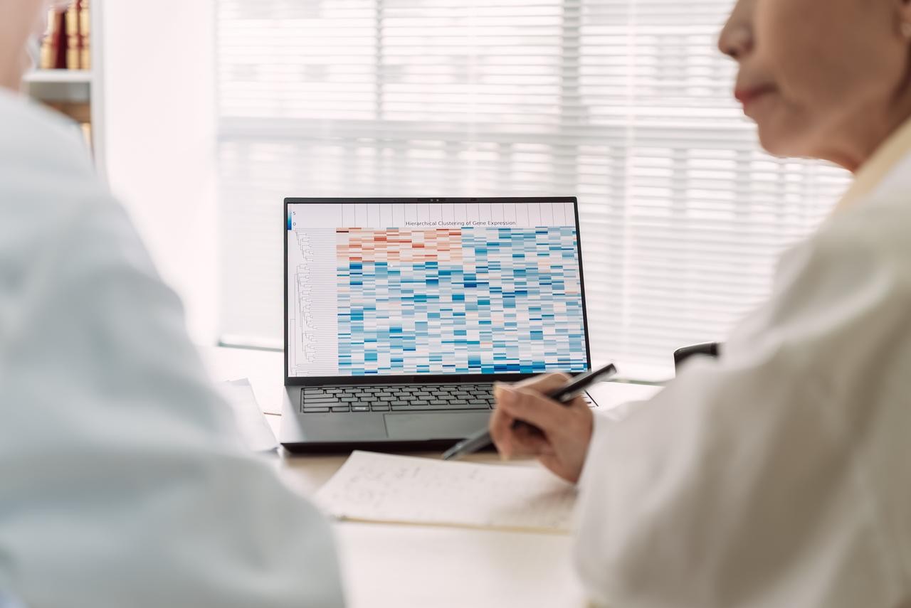 Back and profile close up image of a female HCP holding a pen over a notebook, looking at another HCP with an open laptop screen showing results in the background.