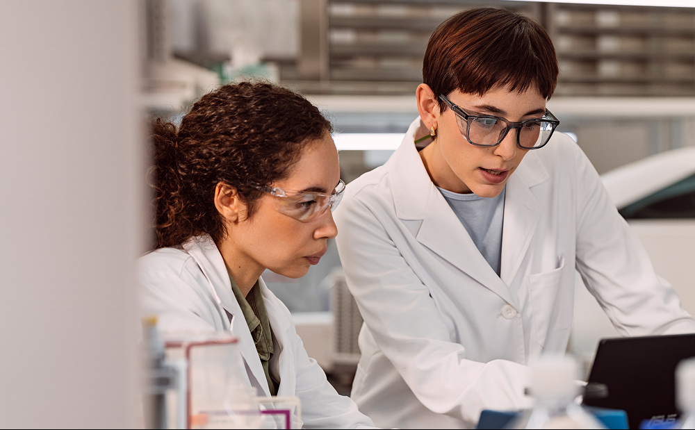 Two female scientists are working together, looking down at a monitor on a laptop in a lab setting.