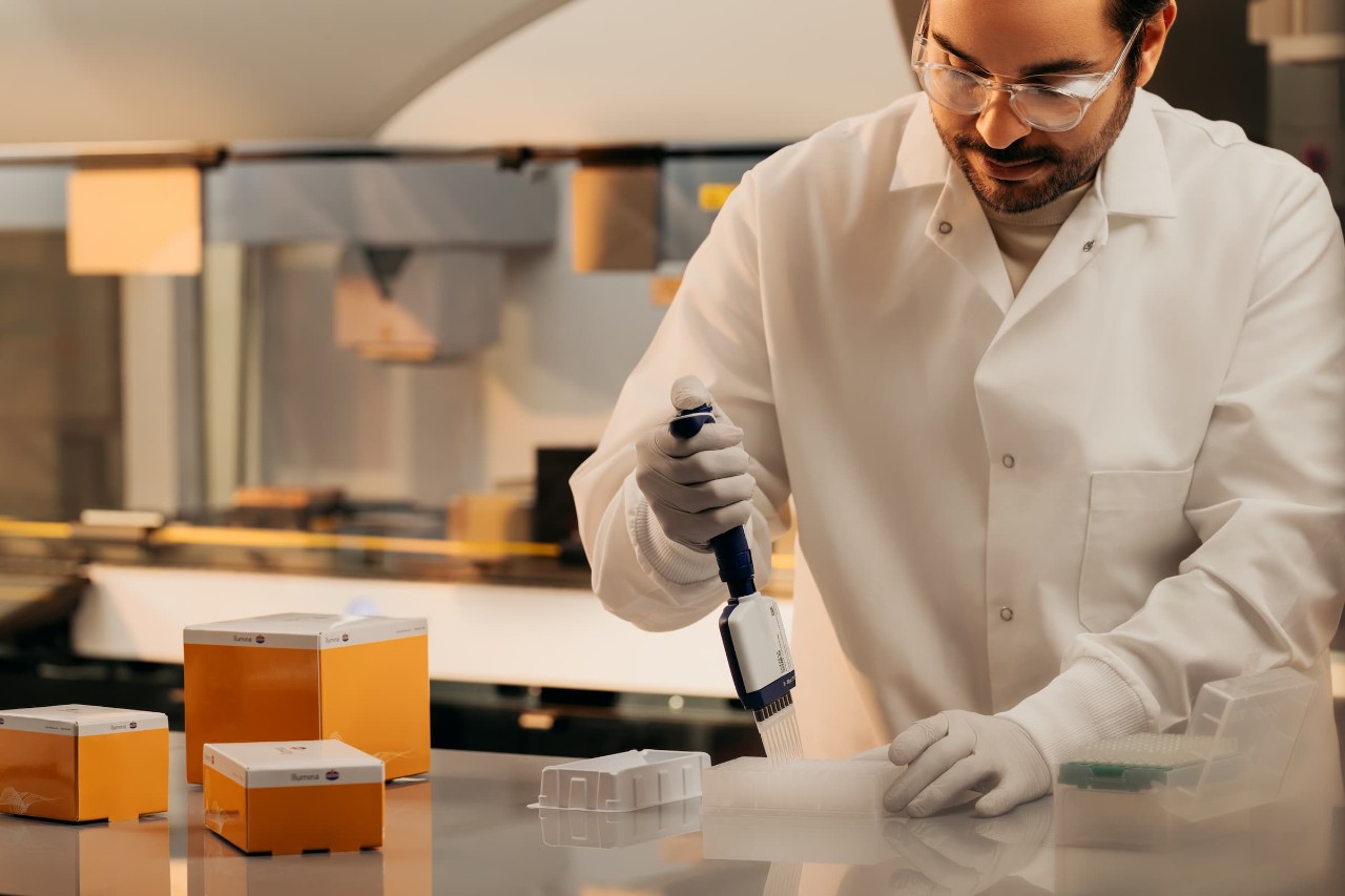 Male scientist is pipetting into a well plate on a lab bench; the Illumina Protein Prep box kit next to him and Tecan workstation in the background