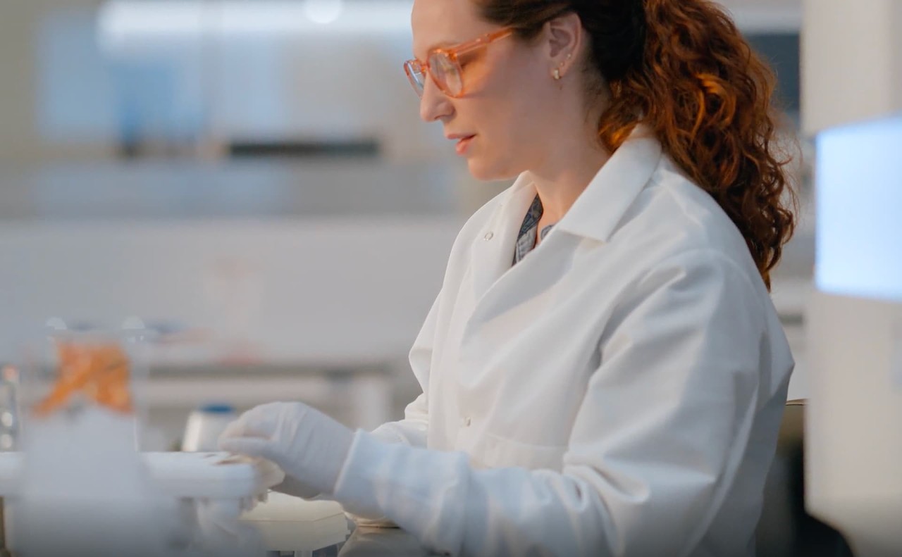 Profile clip of a female scientist placing a library tube strip into a reagent cartridge to prepare for a NovaSeq X run; blurry instrument in foreground.