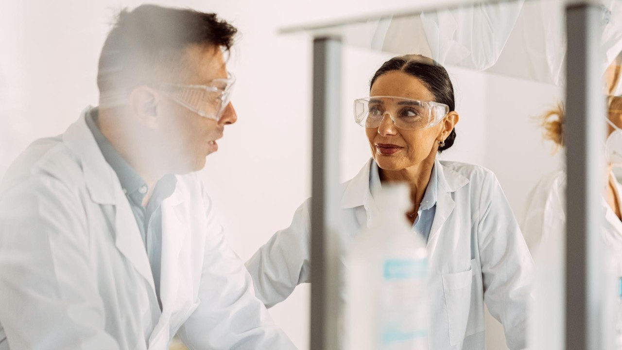Two scientists, one male and one female, engaging in conversation in a lab; tubes and other lab equipment blurry in the foreground.