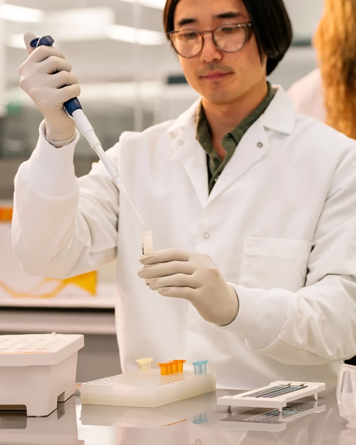 Male scientist holding a library tube in one hand and a single pipette with his other hand, actively pipetting into the tube; flow cell and SBS cartridge on the lab bench in preparation for loading; blurry image of a female scientist interacting with the touch screen monitor on a NovaSeq 6000 and library prep boxes in the background; green status bar